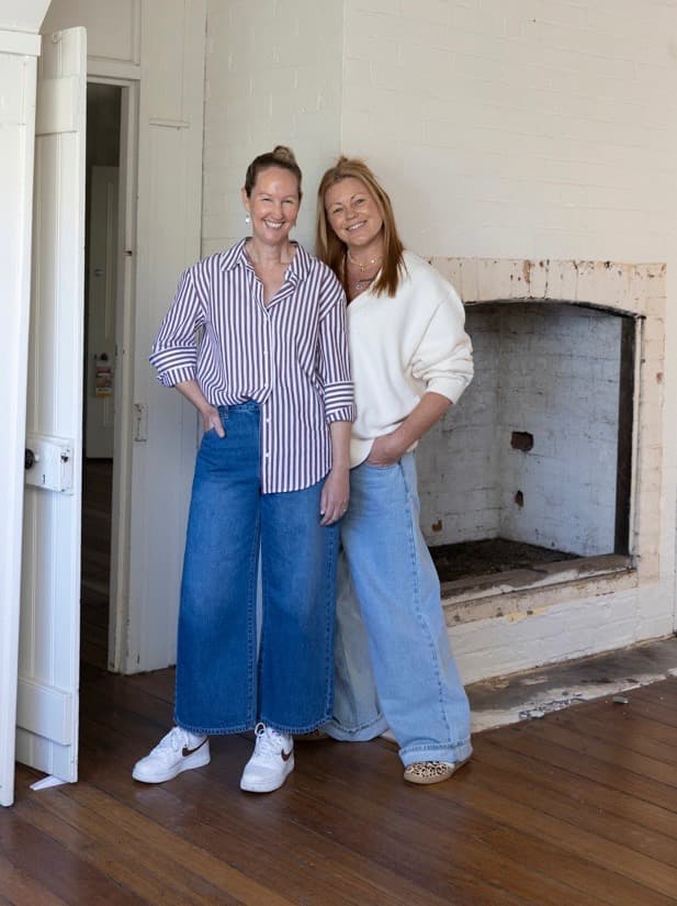 Small Folk founders Bec and Em smiling beside the old brick fireplace inside the heritage cottage